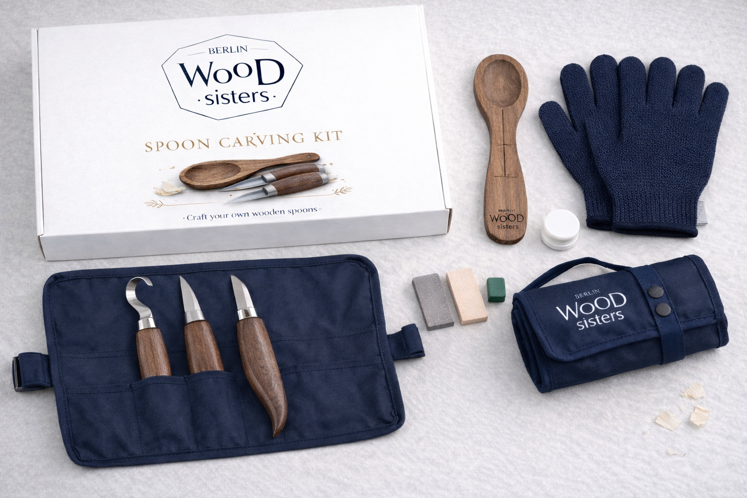 The Berlin Wood Sisters Spoon Carving Kit laid out: presentation box, carving knives in a navy roll, spoon blank, gloves, sharpening stones, honing compound, finishing tin and a branded travel bag
