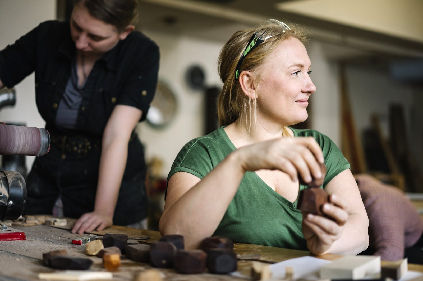 A workshop guest seated with a stack of balancing stones while a teacher sands wood in the background